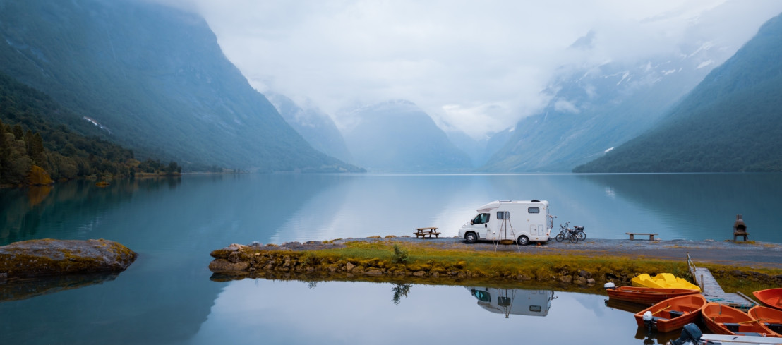 Weißer Camper steht am Ufer eines Fjords, umgeben von steilen Bergen und ruhigem Wasser. Ein spiegelbildähnlicher Effekt im Wasser verstärkt die atemberaubende Landschaft.