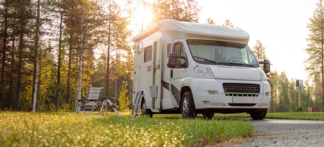 Großer, weißer Camper steht auf einer grünen Wiese auf einem Campingplatz, umgeben von Bäumen.