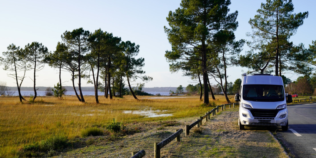 Weißer Camper steht an einer Küstenstraße mit Blick auf einen See und eine natürliche Landschaft mit Kiefern und trockenem Gras.
