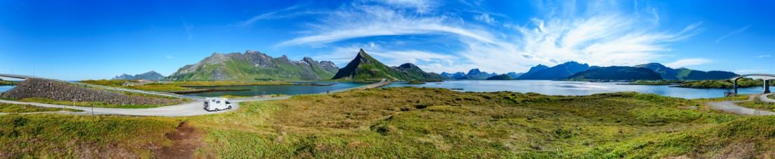 Panoramablick auf einen weißen Camper, der eine Küstenstraße in einer atemberaubenden norwegischen Landschaft entlangfährt. Hohe Berge und tiefblaues Meer im Hintergrund.