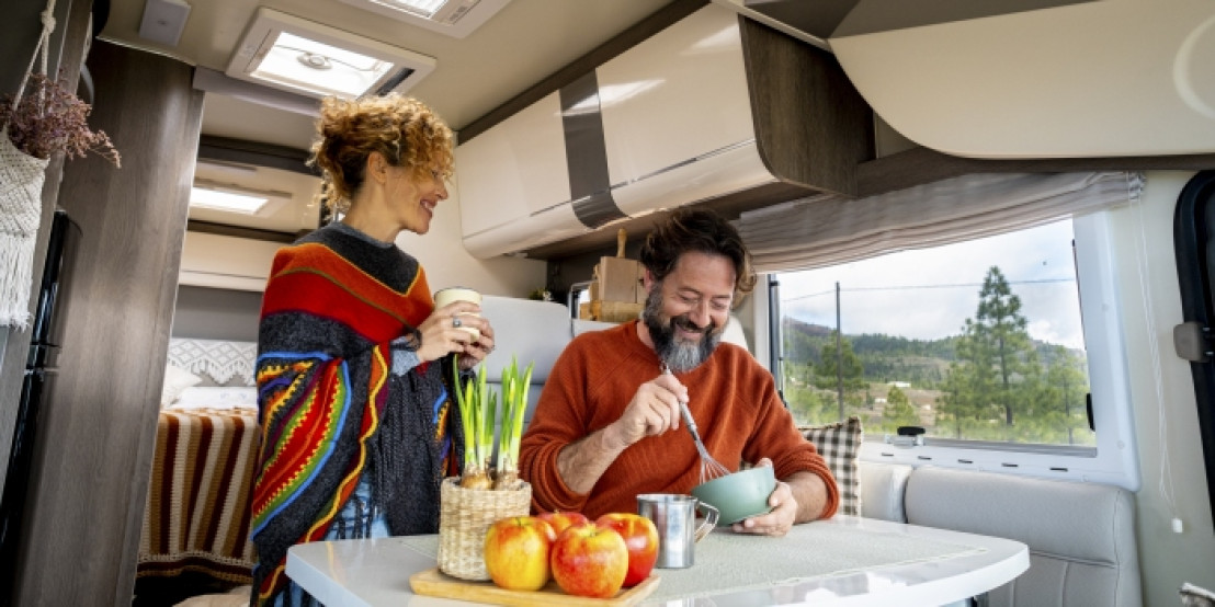 Paar sitzt im Camper an einem Tisch und frühstückt mit Blick auf eine grüne Landschaft.