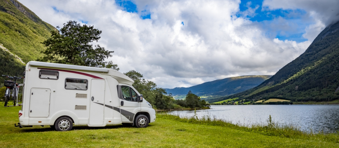 Weißer Camper steht am Ufer eines Sees, umgeben von hohen Bergen und grüner Vegetation in Norwegen.