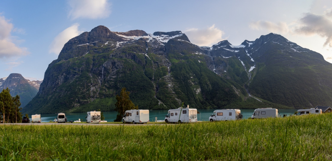 Mehrere Camper stehen auf einer Wiese vor hohen, grünen Bergen und einem tiefblauen See