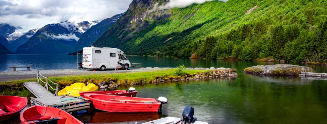 Weißer Camper steht direkt am Ufer eines Fjords, umgeben von hohen Bergen und grüner Vegetation. Rote Ruderboote liegen im Vordergrund an einem Steg. Traumhafte Szenerie für einen Campingurlaub in der Natur.