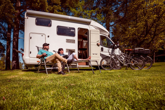 Ein Paar entspannt sich in Liegestühlen vor ihrem Camper auf einer grünen Wiese. Im Hintergrund stehen Fahrräder bereit für Ausflüge.