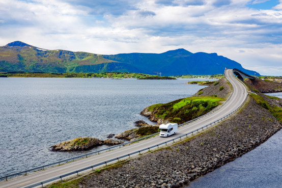 Weißer Camper fährt auf einer Küstenstraße durch eine spektakuläre Landschaft mit Bergen und Meer.