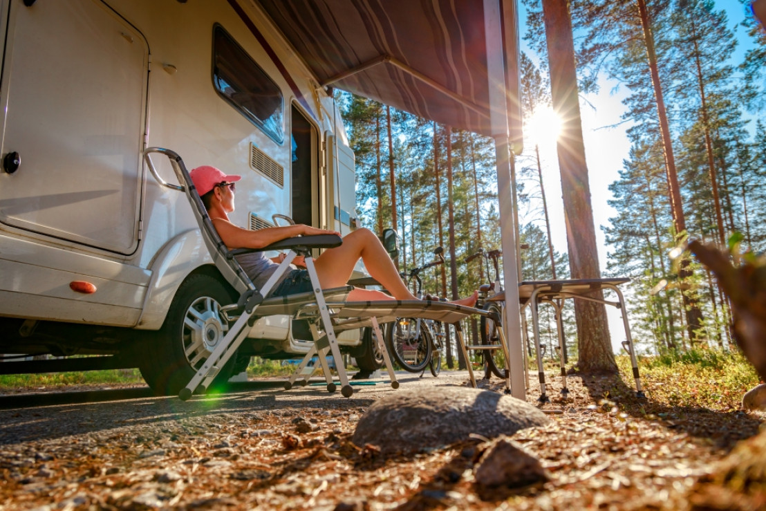 Eine Frau sitzt vor ihrem Wohnmobil auf einem Campingplatz inmitten waldreicher Natur.